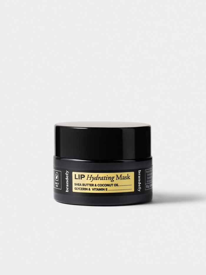 The Lip Hydrating Mask in a matte black jar with a gold-yellow label, on a light grey ground.