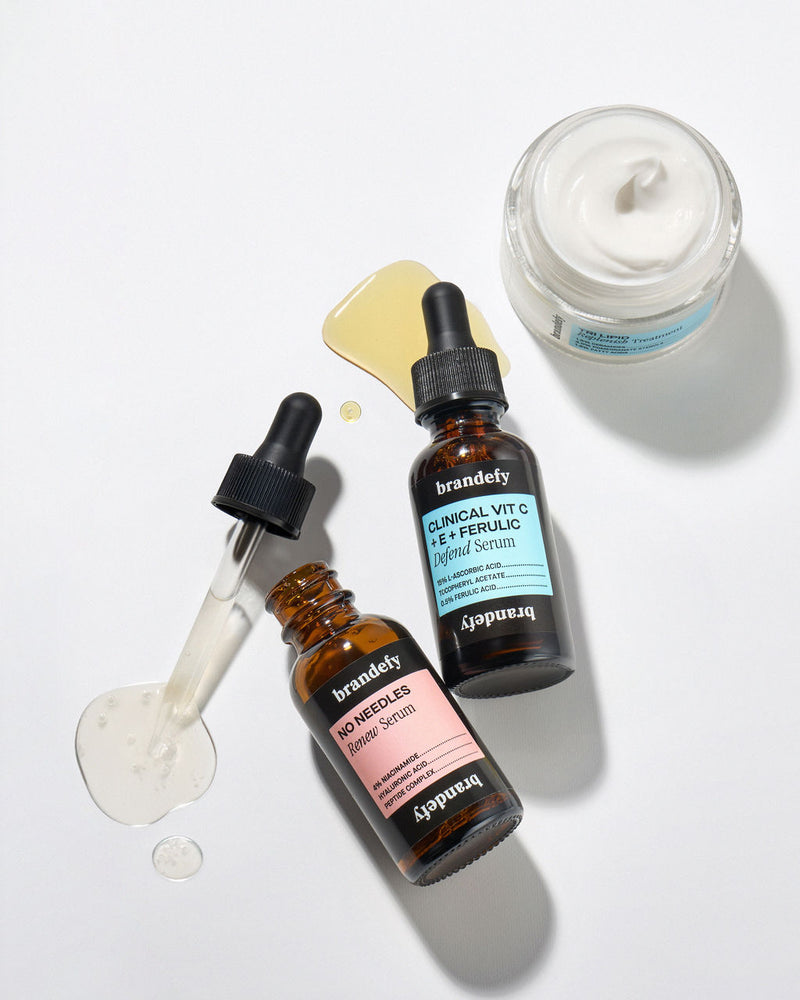 Overhead flat lay of three Brandefy products with texture swatches: the amber Vit C + E + Ferulic Defend Serum, the No Needles Renew Serum, and an open jar of Tri Lipid Replenish Treatment.