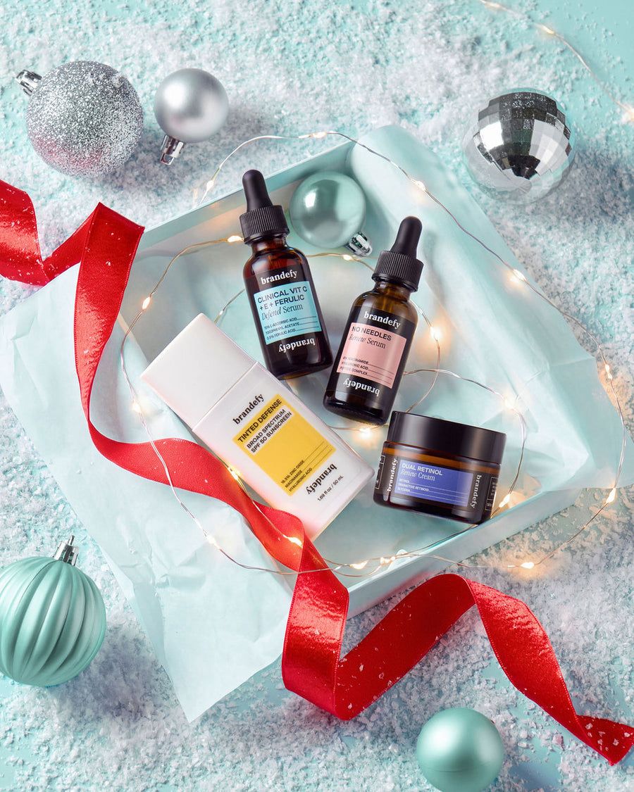 Flat-lay of the four products in The 2 Minute Method arranged in a pale mint tray with fairy lights, a red ribbon and silver and mint baubles on a snowy ground: the Clinical Vit C + E + Ferulic Defend Serum and No Needles Renew Serum in amber dropper bottles, the white Tinted Defense SPF 50 tube, and the Dual Retinol Renew Cream jar.