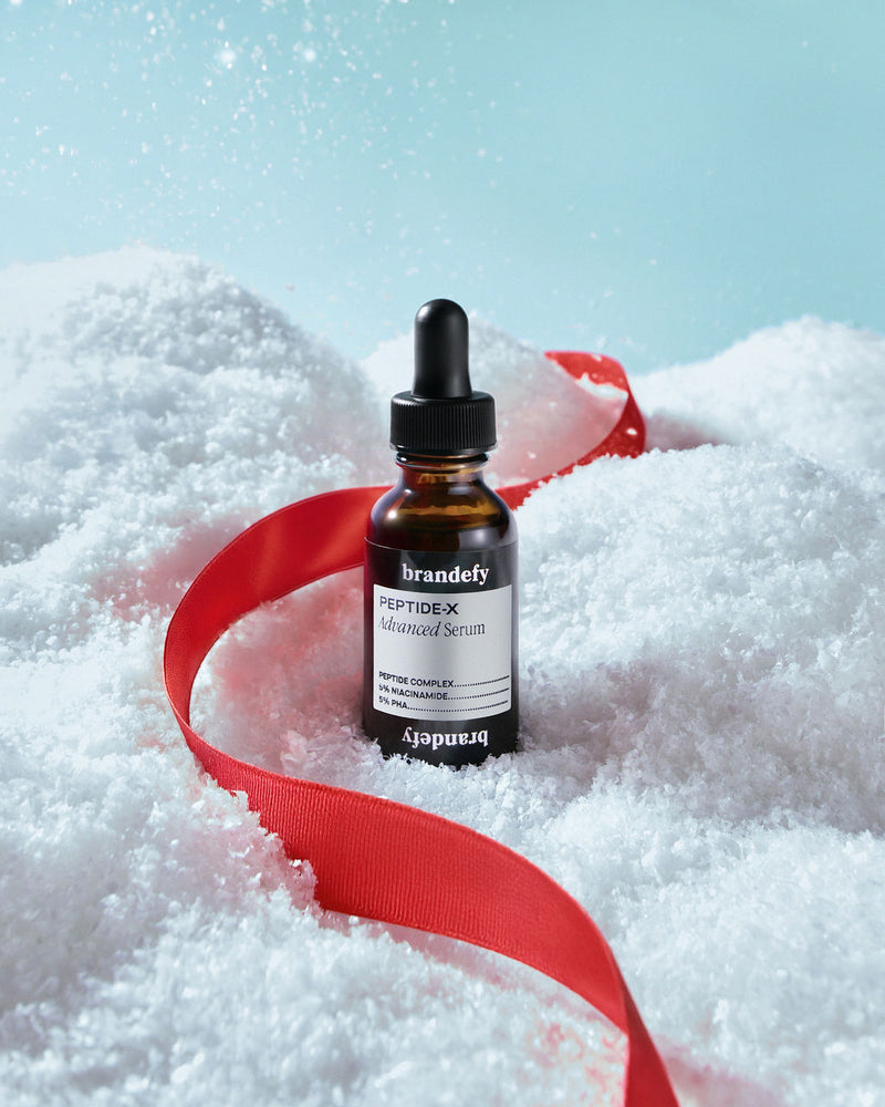 The Peptide-X Advanced Serum bottle standing in fresh snow with a red satin ribbon curling around it.