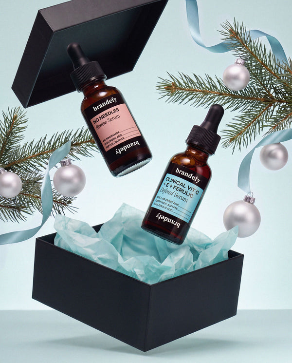The No Needles Renew Serum and the Clinical Vit C + E + Ferulic serum above an open black gift box, with pine sprigs and silver baubles.
