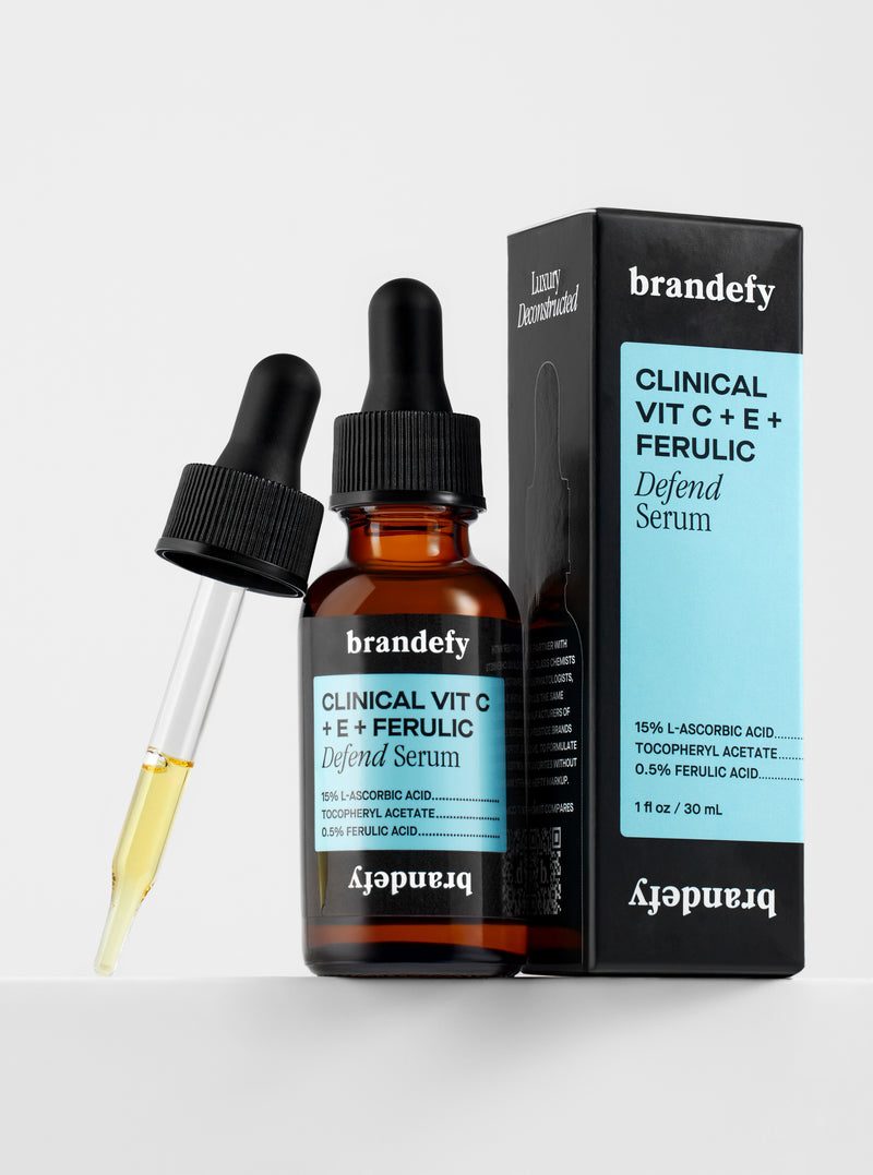 Brandefy Clinical Vit C + E + Ferulic Defend Serum: an amber glass dropper bottle with a light blue label reading 15% L-ascorbic acid, its glass dropper wand filled with pale yellow serum resting against it, beside the black retail carton.