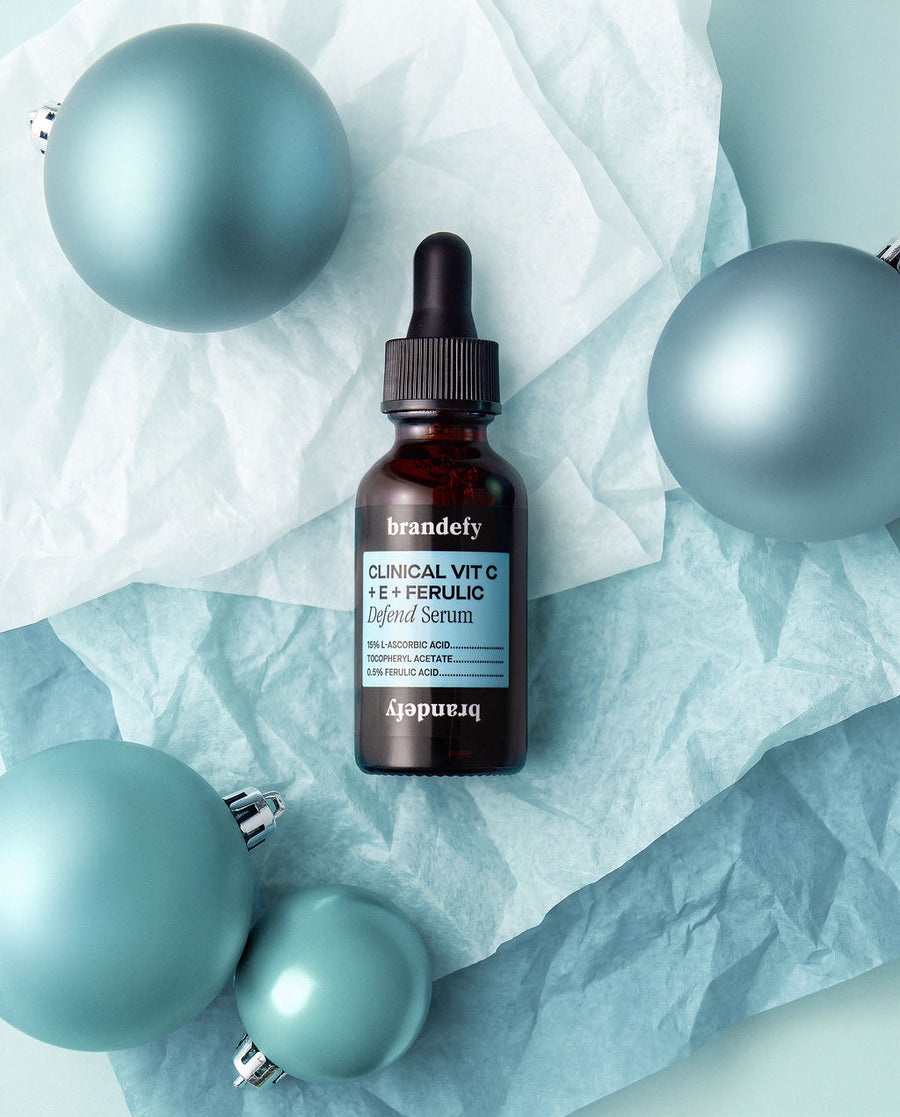 The Clinical Vit C + E + Ferulic Defend Serum bottle on pale tissue paper, surrounded by matte teal Christmas baubles.