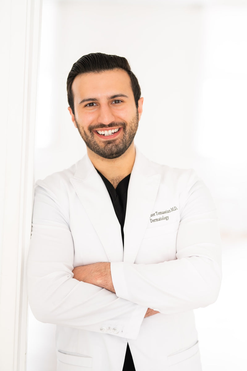Dr. Chris Tomassian, dermatologist, in a white lab coat, arms folded, in a bright white room.