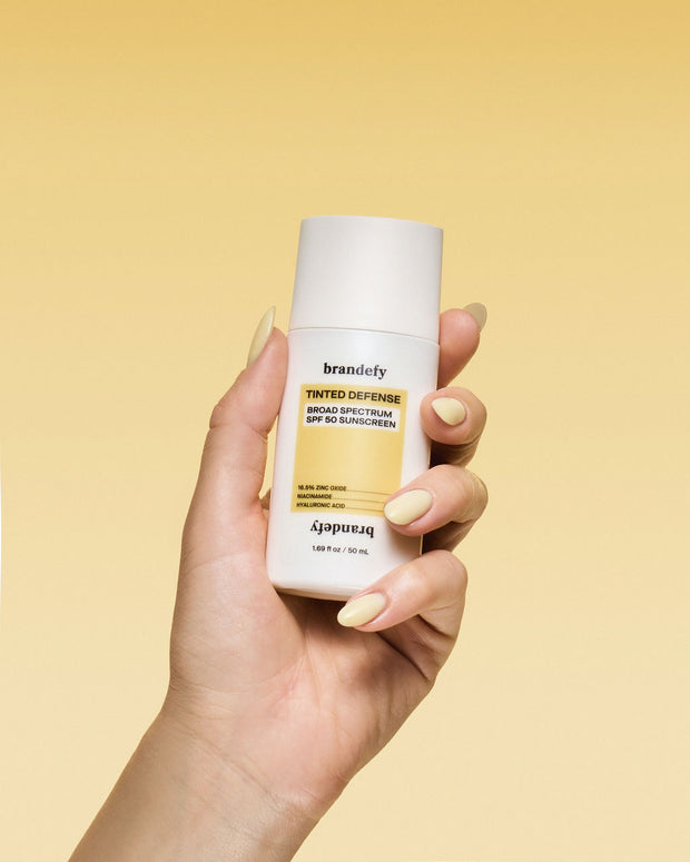 A hand holds a white bottle of Brandefy Tinted Defense Broad Spectrum SPF 50 Sunscreen, yellow label, against a plain yellow background.