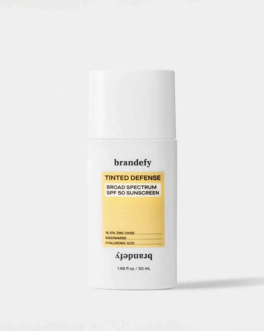 A white bottle of Brandefy Tinted Defense Broad Spectrum SPF 50 Sunscreen with a yellow label reading 16.5% zinc oxide, niacinamide and hyaluronic acid.