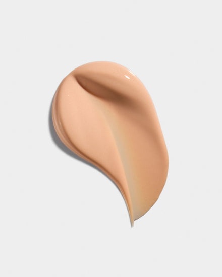 A swatch of the Tinted Defense sunscreen: a glossy, warm peach-beige cream swirled into a comma shape on a white background.